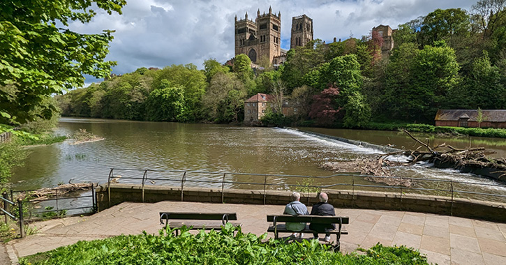 couple sat on a bench overlooking the River Wear and with Durham Cathedral in the background on a sunny day.
